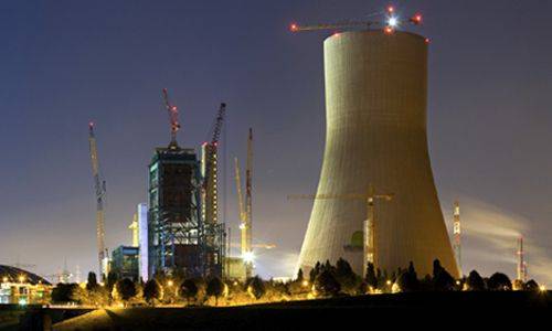 An industrial site under construction at night featuring a tall cooling tower and several cranes. The cooling tower is illuminated, with a partially completed building nearby surrounded by construction equipment and lights.